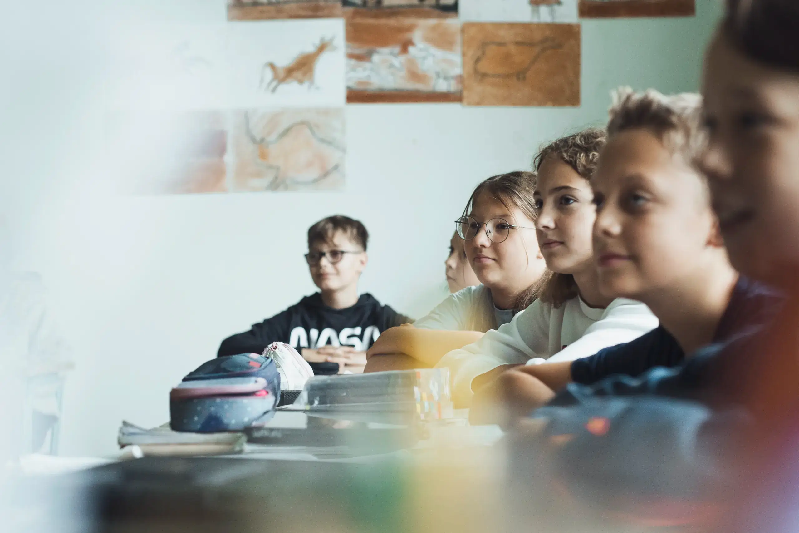 HebbelCampus Eider Nordsee Schule Unterricht2 Mehrere Kinder sitzen an einem Tisch in einem Klassenzimmer und schauen nach vorne. Im Hintergrund sind Zeichnungen an der Wand zu sehen. Auf dem Tisch liegen Schulutensilien. Das Setting wirkt konzentriert und ruhig.