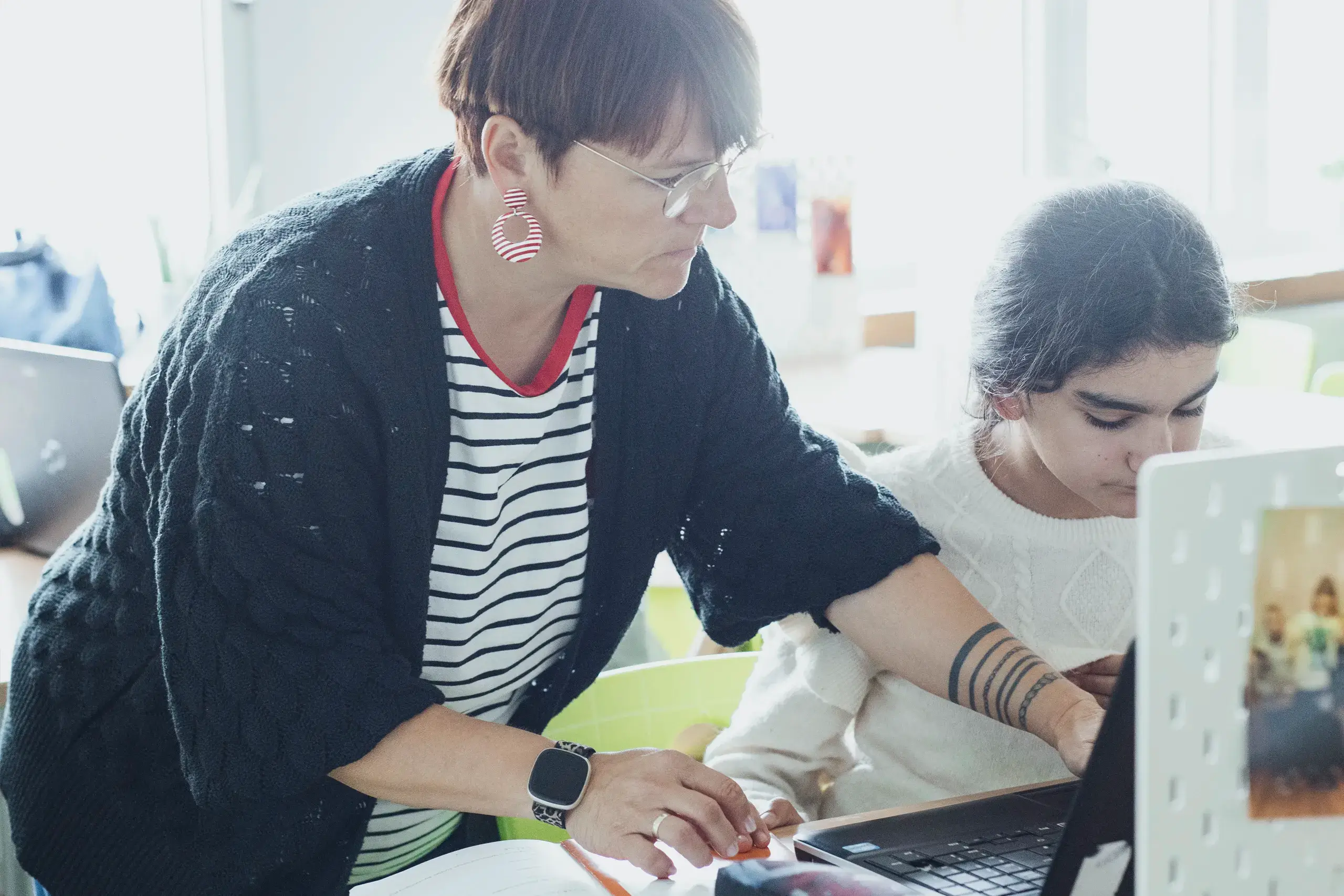 HebbelCampus Eider Nordsee Schule Unterricht3 Eine Person mit Brille und gestreiftem Shirt hilft einer anderen Person bei der Arbeit am Laptop. Beide sind in einem gut beleuchteten Raum mit mehreren Computern.