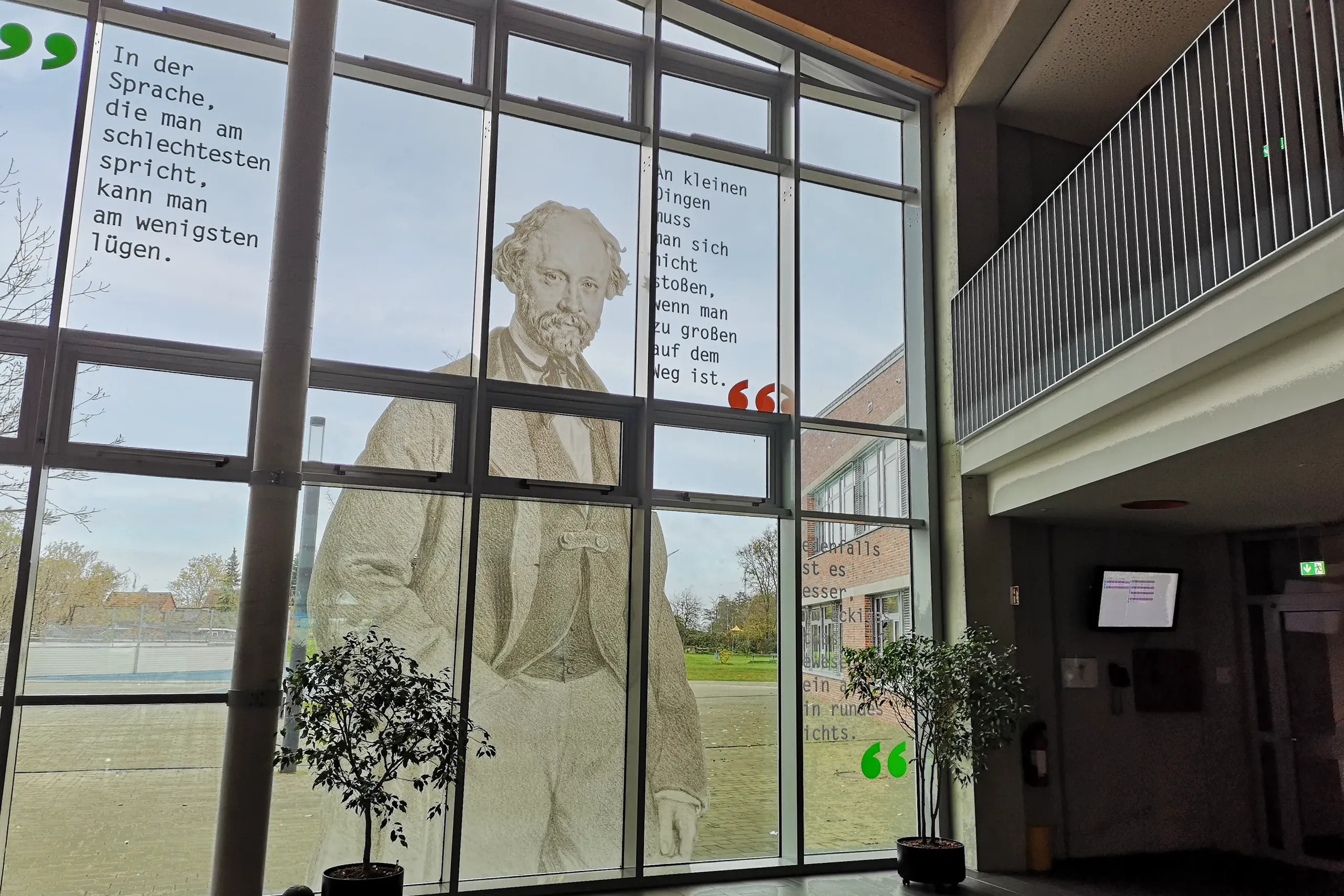 Ein großes Fenster in einem Gebäude zeigt das Bild eines Mannes in historischer Kleidung. Auf dem Fenster sind Zitate in verschiedenen Farben gedruckt. Im Vordergrund stehen zwei kleine Pflanzen in Töpfen.