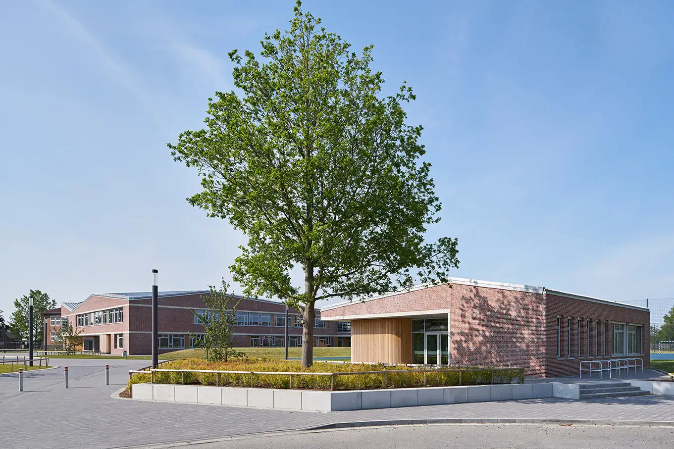 Hebbel Campus ENS Wesselburen Ein moderner Schulkomplex aus rotem Backstein bei sonnigem Wetter. Im Vordergrund steht ein Baum in einem bepflanzten Bereich, umgeben von asphaltierten Wegen und einem niedrigen Zaun. Im Hintergrund sieht man ein größeres Gebäude.