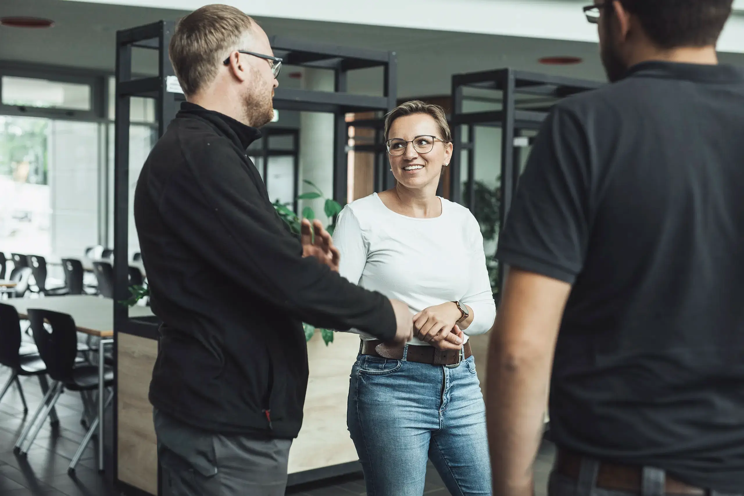 Eine Gruppe von drei Personen steht in einem modernen Büro oder Café und unterhält sich. Eine Person trägt eine Brille und lächelt. Im Hintergrund sind Stühle, Tische und Pflanzen zu sehen.