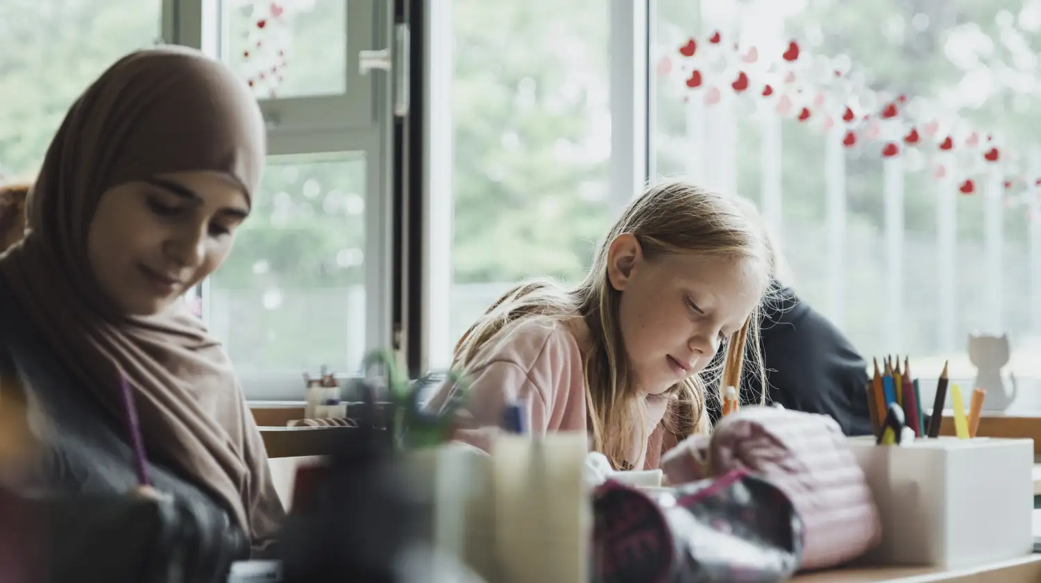 Hebbel Campus Dummy Wer Wir Sind Zwei Personen sitzen in einem Klassenraum vor einem Fenster. Eine Person trägt ein Kopftuch, die andere hat langes blondes Haar. Beide arbeiten konzentriert an einem Tisch mit Schulsachen. Im Hintergrund sind Fensterdekorationen zu sehen.