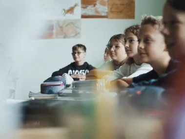 Eine Gruppe von Kindern sitzt gemeinsam an einem Tisch in einem Klassenzimmer. Im Hintergrund sind Bilder von Tieren und Landschaften an der Wand zu sehen. Der Raum ist hell beleuchtet.