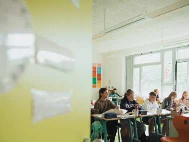 In einem hellen Klassenzimmer sitzen mehrere Schülerinnen und Schüler an Tischen und arbeiten. Eine gelbe Tür steht offen. Im Hintergrund sind große Fenster und bunte Plakate an der Wand zu sehen.