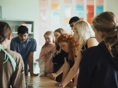 Eine Gruppe von jungen Menschen steht dicht gedrängt um einen Tisch und betrachtet etwas darauf. Im Hintergrund sind bunte Poster an der Wand. Die Personen wirken konzentriert und diskutierend.