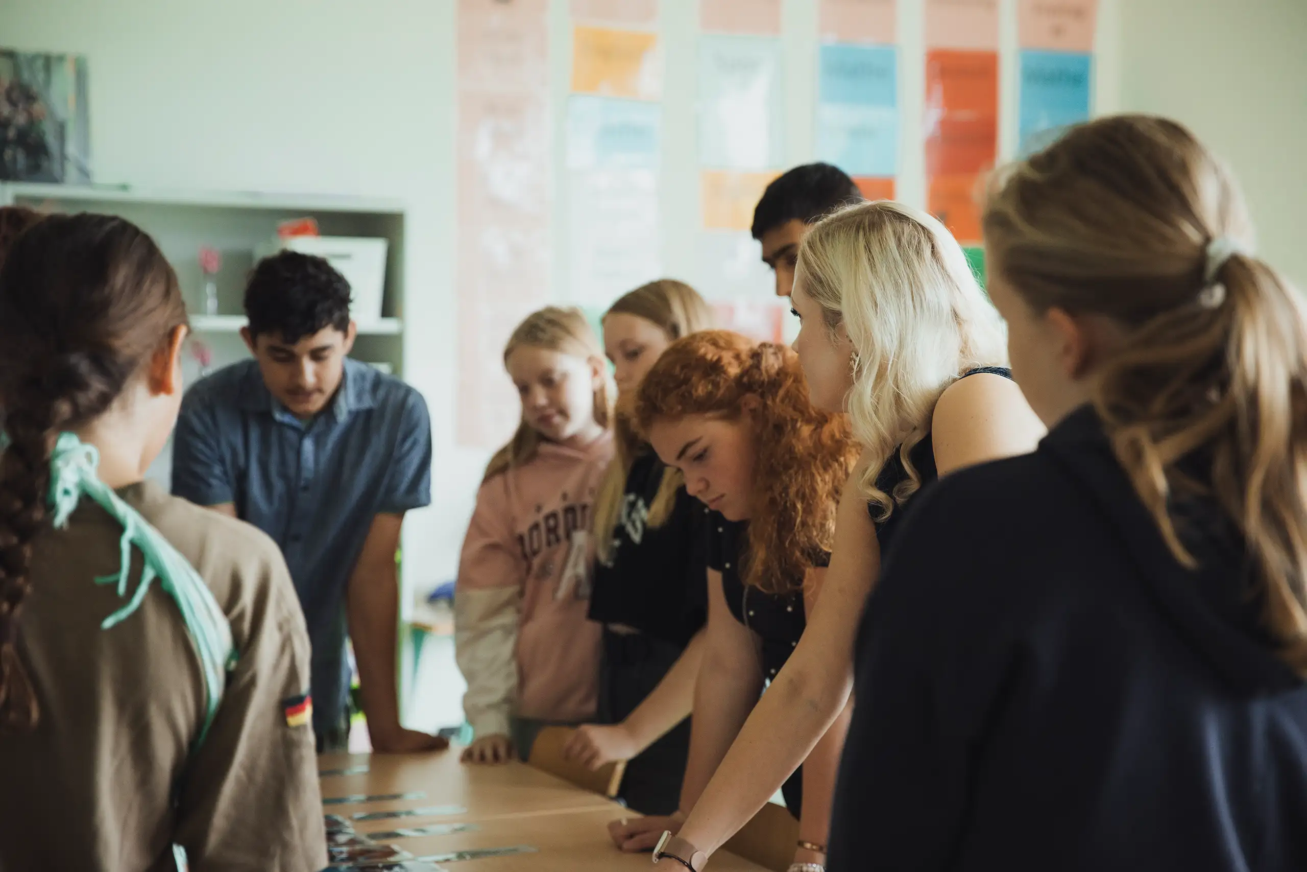 Eine Gruppe von jungen Menschen steht dicht gedrängt um einen Tisch und betrachtet etwas darauf. Im Hintergrund sind bunte Poster an der Wand. Die Personen wirken konzentriert und diskutierend.