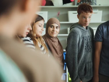 Eine Gruppe von Jugendlichen steht in einer Reihe in einem Raum mit Regalen. Die Personen stehen nah beieinander und scheinen in ein Gespräch vertieft zu sein. Eine Person in der Mitte trägt ein Kopftuch.