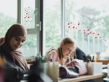 Eine Person mit Kopftuch und ein Kind sitzen in einem Klassenzimmer an einem Tisch und schreiben. Im Hintergrund sind Fenster mit roten Herzdekorationen. Der Tisch ist mit Stiften und Federmäppchen bestückt.