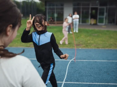 Ein Kind in einem schwarz-blauen Trainingsanzug zeigt ein Peace-Zeichen auf einem blauen Sportplatz. Ein anderes Kind steht ihm gegenüber. Im Hintergrund spielen weitere Kinder nahe einem Gebäude.