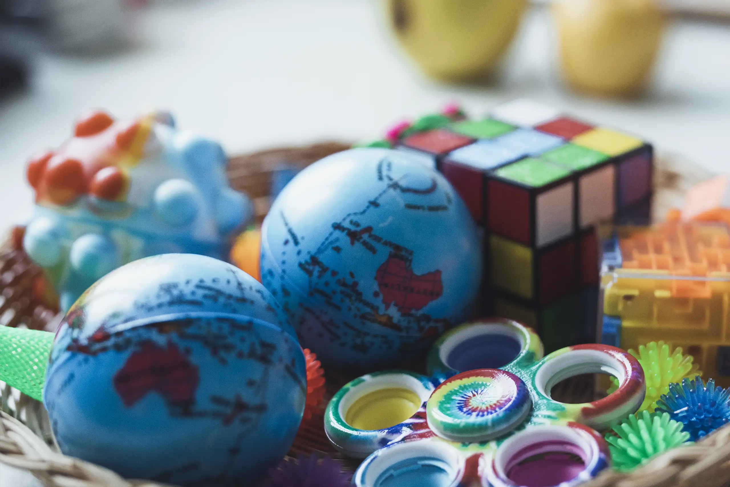 Ein Korb voller bunter Spielzeuge: Zwei blaue Erdkugel-Stressbälle mit Weltkartendruck, ein bunter Zauberwürfel, ein blau-orangefarbenes Noppenballspielzeug und mehrere bunte Spiralspielzeuge.