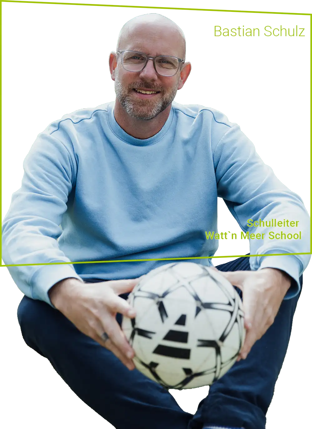 Hebbel Campus Team Bastian Schulz Ein Mann mit Brille und Bart sitzt auf dem Boden und hält einen Fußball. Er trägt einen hellblauen Pullover. Im Hintergrund ist eine unscharfe grüne Fläche zu sehen. Text: "Schulleiter Watt'n Meer School".