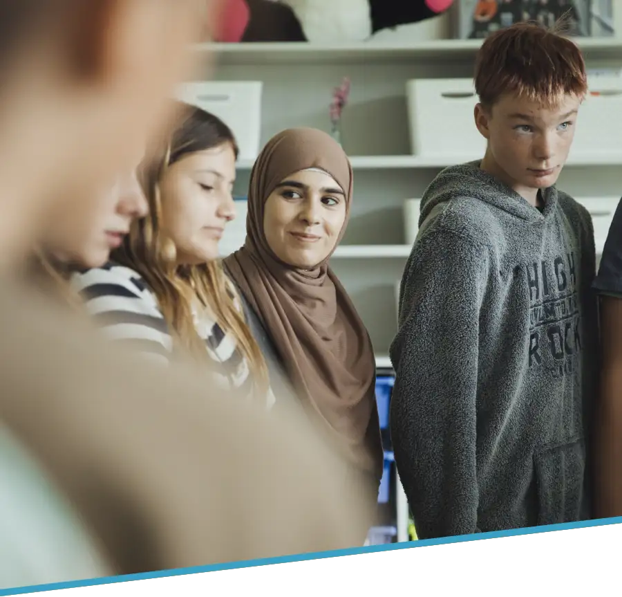 Hebbel Campus Teaser Jugendliche 1 Eine Gruppe von Jugendlichen steht in einem Kreis und blickt in die Mitte. Sie befinden sich in einem Raum mit Regalen und Kisten im Hintergrund. Die Stimmung wirkt aufmerksam und freundlich.
