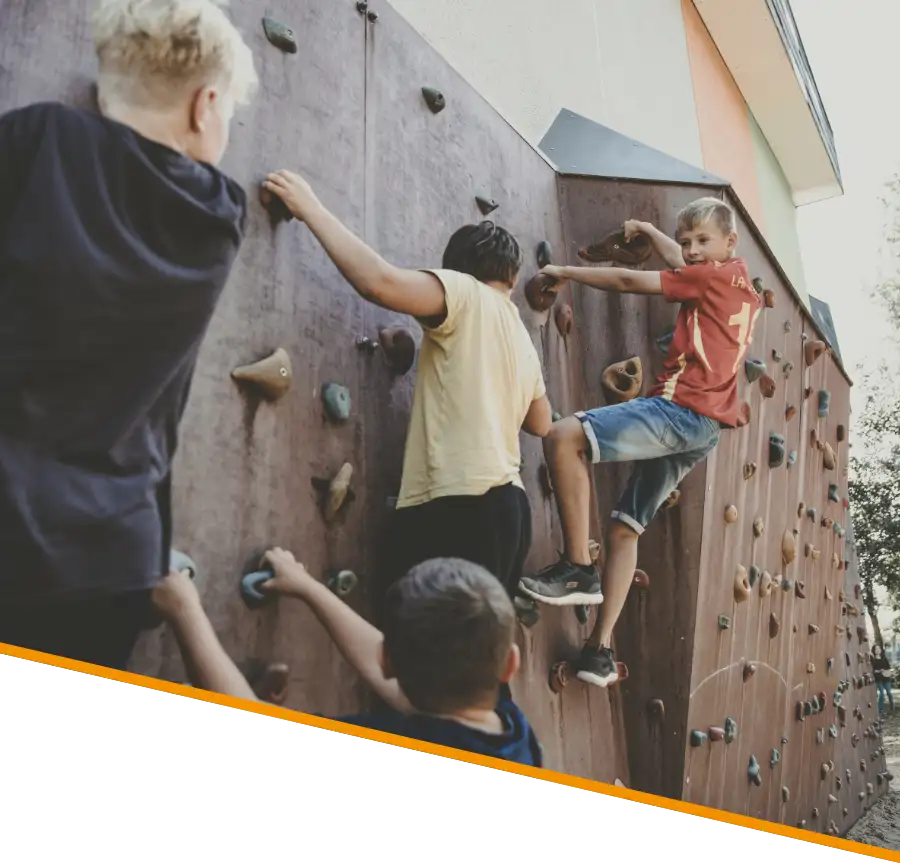 Hebbel Campus Teaser Kinder 1 Eine Gruppe von Kindern klettert an einer Kletterwand im Freien. Die Wand hat bunte Griffe und die Kinder tragen Freizeitkleidung. Im Hintergrund sind Bäume und ein Gebäude zu sehen.