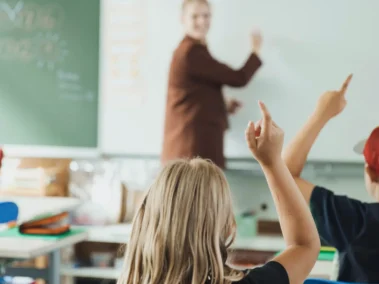 Eine Lehrerin schreibt an eine Tafel, während Kinder im Vordergrund mit erhobenen Händen sitzen. Die Szene spielt in einem Klassenzimmer mit Tischen, Stühlen und Schulmaterialien. Die Stimmung wirkt engagiert und lernfreudig.