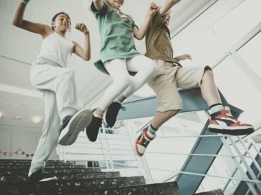 Drei Kinder springen freudig eine Treppe in einem modernen Gebäude hinunter. Sie tragen lässige Alltagskleidung und Turnschuhe. Die Szene wirkt dynamisch und fröhlich.