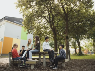 Eine Gruppe von sechs Jugendlichen sitzt auf und um einen Picknicktisch im Freien. Sie unterhalten sich und lachen, umgeben von Bäumen und einem Gebäude mit bunten Wänden im Hintergrund. Die Sonne scheint.