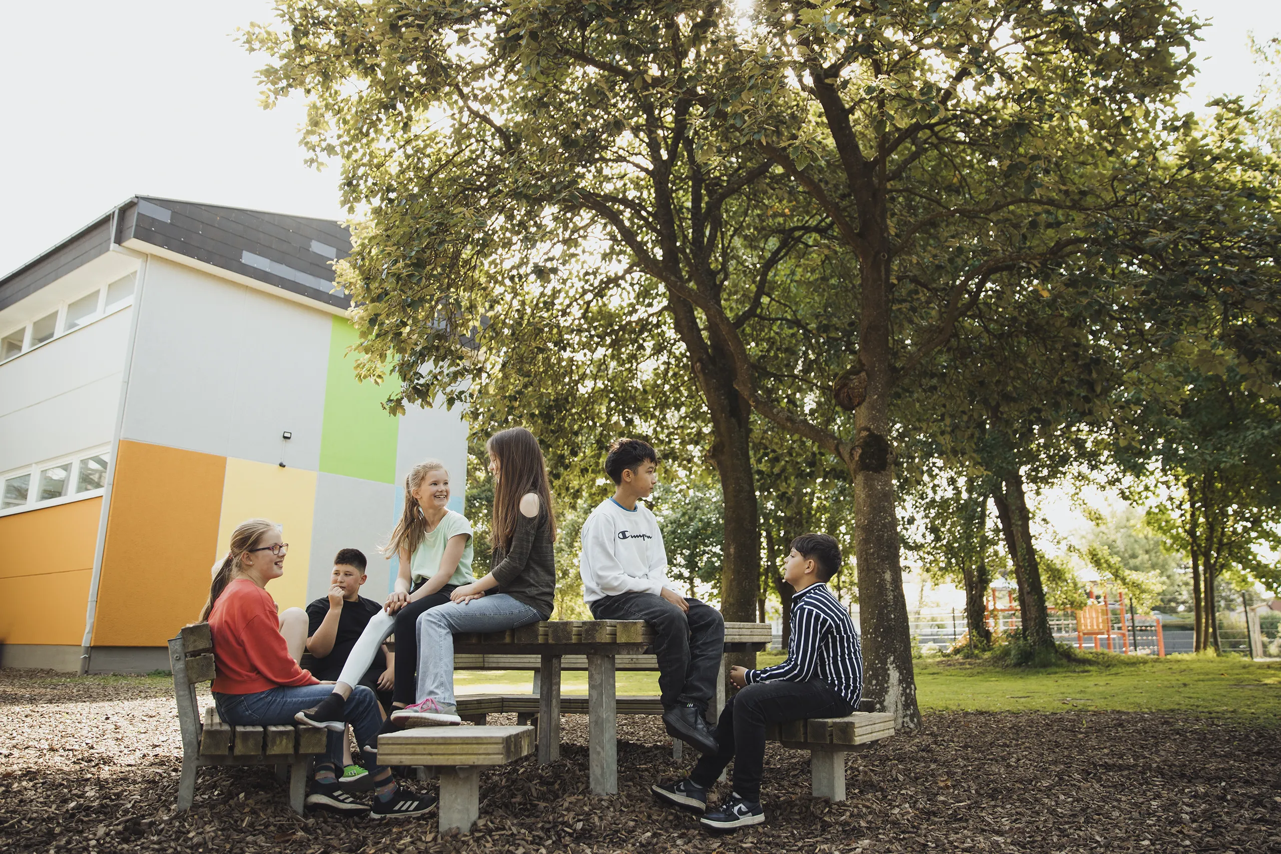 Eine Gruppe von sechs Jugendlichen sitzt auf und um einen Picknicktisch im Freien. Sie unterhalten sich und lachen, umgeben von Bäumen und einem Gebäude mit bunten Wänden im Hintergrund. Die Sonne scheint.