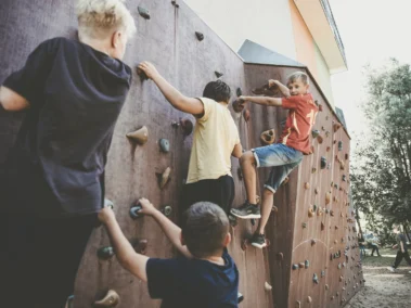 Vier Kinder klettern an einer Kletterwand im Freien. Die Wand hat verschiedene bunte Griffe. Es ist ein sonniger Tag und im Hintergrund sind Bäume und weiteres Gelände eines Spielplatzes zu sehen.