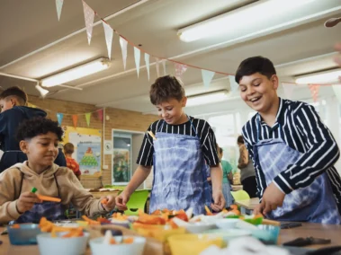 Kinder schneiden buntes Gemüse in einem hellen Raum mit Partydekorationen. Drei Jungen im Vordergrund tragen gestreifte Kleidung und bunte Schürzen. Weitere Kinder sind im Hintergrund beschäftigt. Tische sind mit Schalen und Bastelutensilien gefüllt.