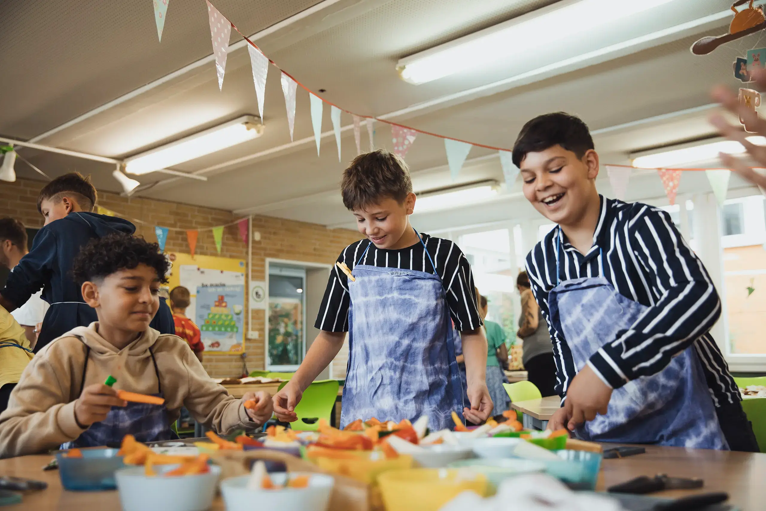Hebbel Campus Wattn Meer School Kueche Kinder schneiden buntes Gemüse in einem hellen Raum mit Partydekorationen. Drei Jungen im Vordergrund tragen gestreifte Kleidung und bunte Schürzen. Weitere Kinder sind im Hintergrund beschäftigt. Tische sind mit Schalen und Bastelutensilien gefüllt.