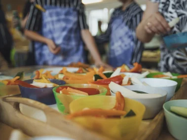 Verschiedene bunte Schalen mit geschnittenem Gemüse wie Paprika und Gurken stehen auf einem Tisch. Menschen im Hintergrund tragen bunte Schürzen und greifen nach dem Essen. Die Szene wirkt lebendig und fröhlich.