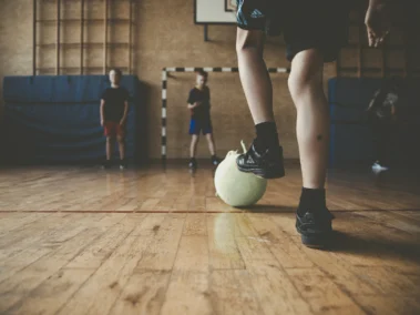 Ein paar junge Menschen spielen in einer Sporthalle Fußball. Im Vordergrund ist der Unterschenkel einer Person zu sehen, die einen Ball mit dem Fuß führt. Weitere Personen stehen im Hintergrund bei einem Tor.