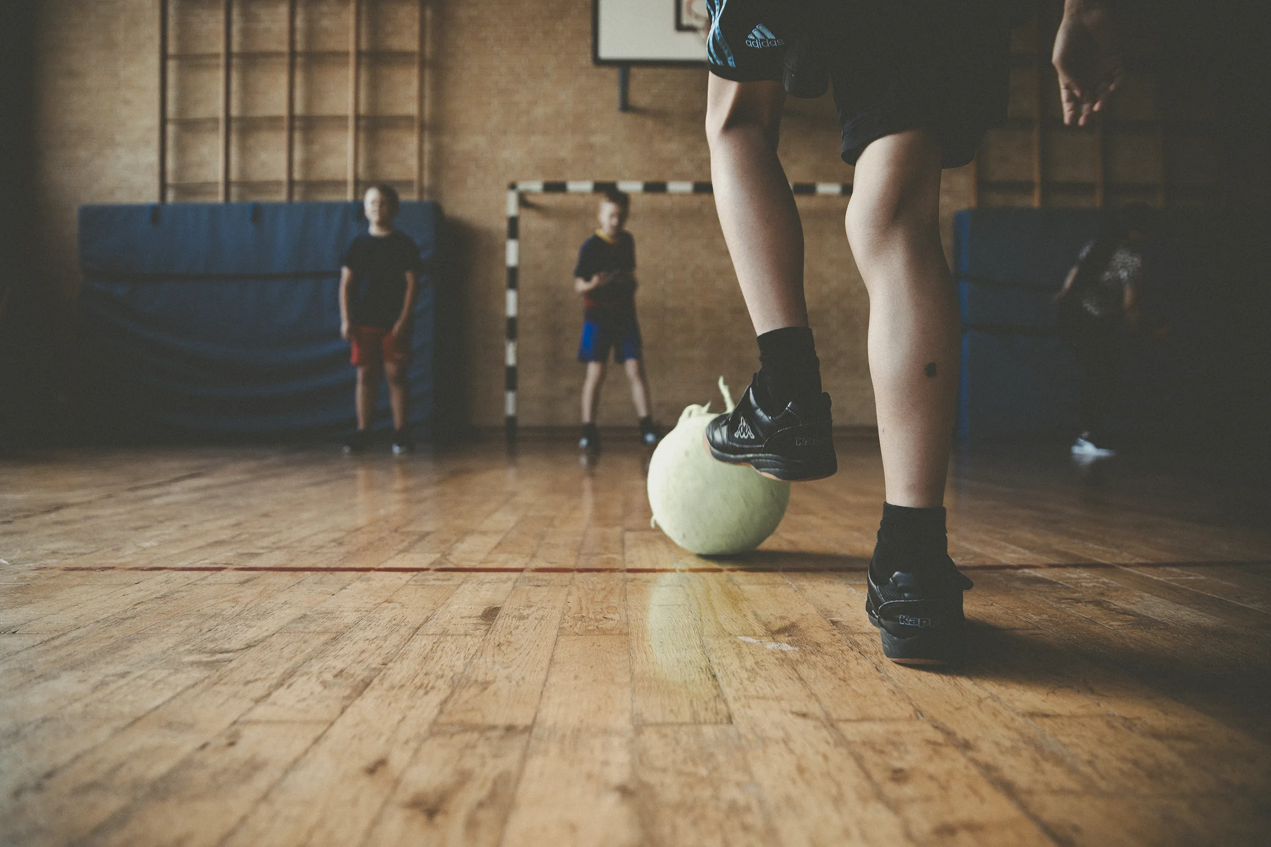 Ein paar junge Menschen spielen in einer Sporthalle Fußball. Im Vordergrund ist der Unterschenkel einer Person zu sehen, die einen Ball mit dem Fuß führt. Weitere Personen stehen im Hintergrund bei einem Tor.