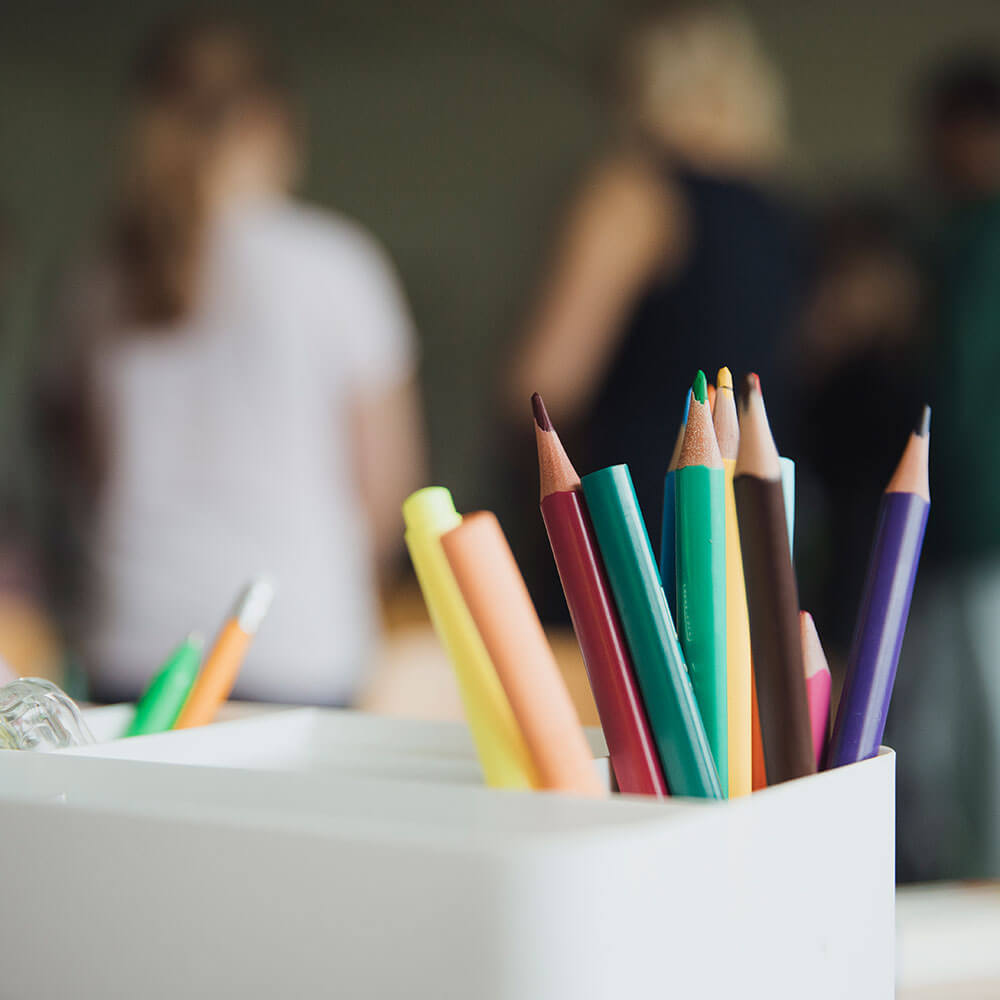 Hebbel Dummy 2 Ein Behälter mit bunten Bleistiften und Stiften steht im Vordergrund. Im Hintergrund sind unscharf Personen zu sehen, die sich unterhalten oder arbeiten.