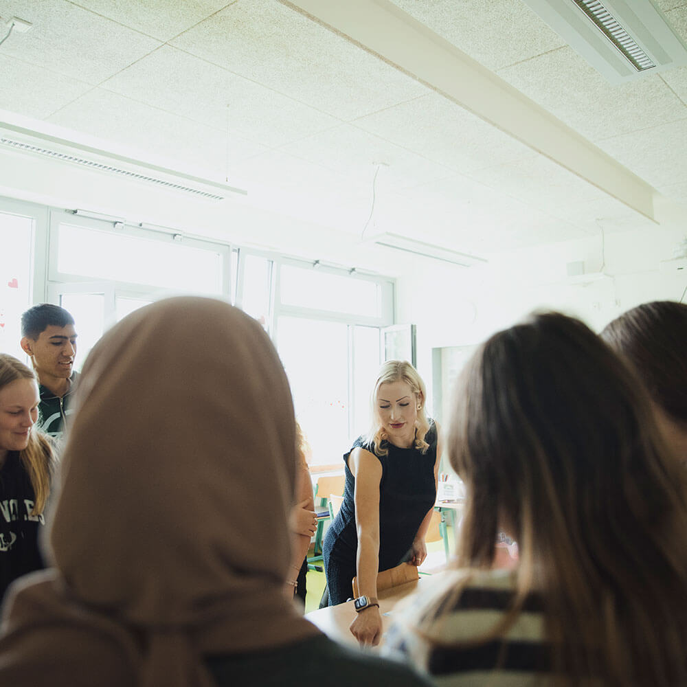 Hebbel Dummy 3 Ein Gruppen von Personen steht um einen Tisch in einem hellen Raum. Eine Person zeigt auf etwas auf dem Tisch. An den Wänden sind große Fenster, durch die Tageslicht einfällt.